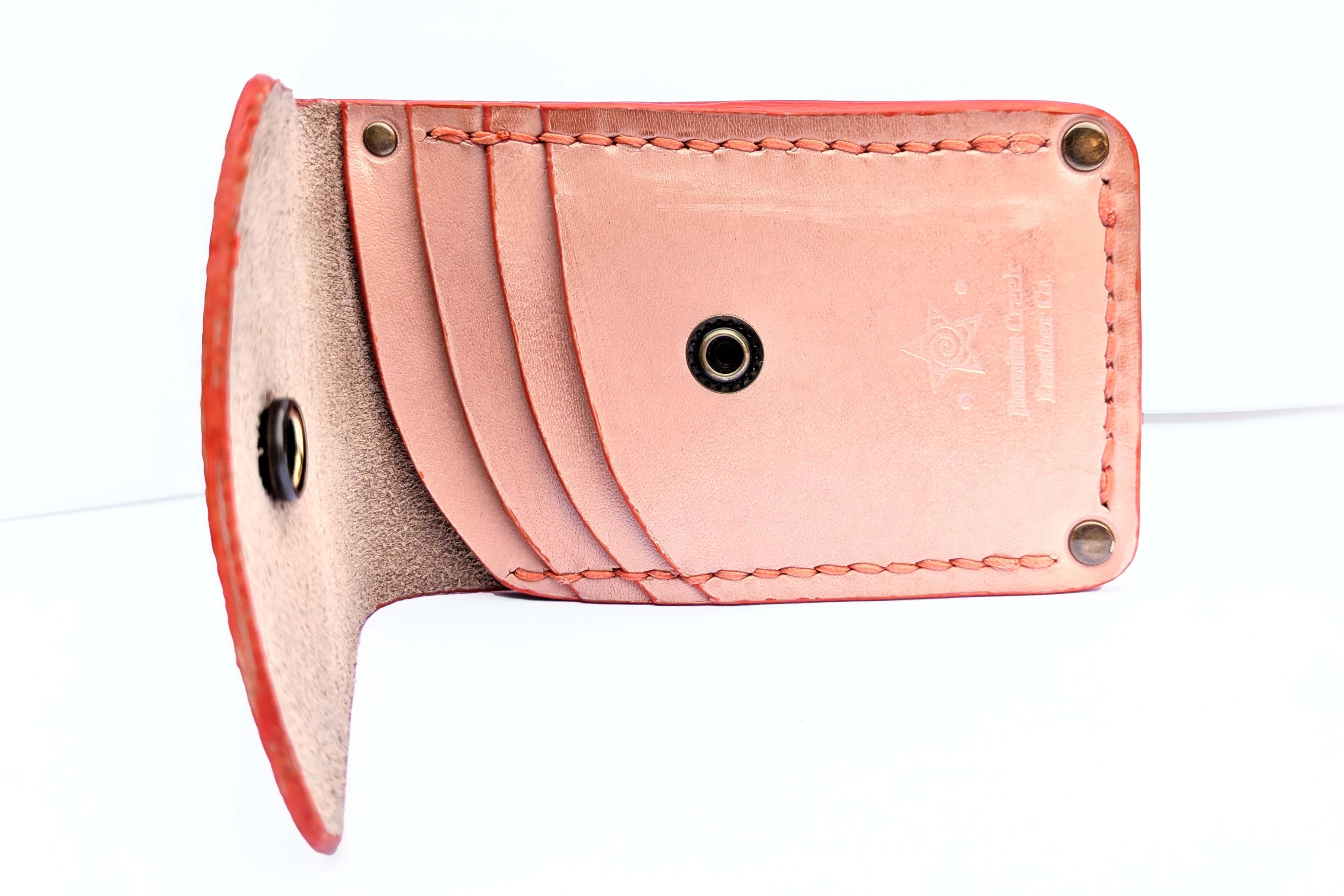 A close-up of a light pinkish-beige leather card holder or wallet shown open, with multiple card slots, visible stitching, brass-tone snaps, and a small embossed logo on a white background.