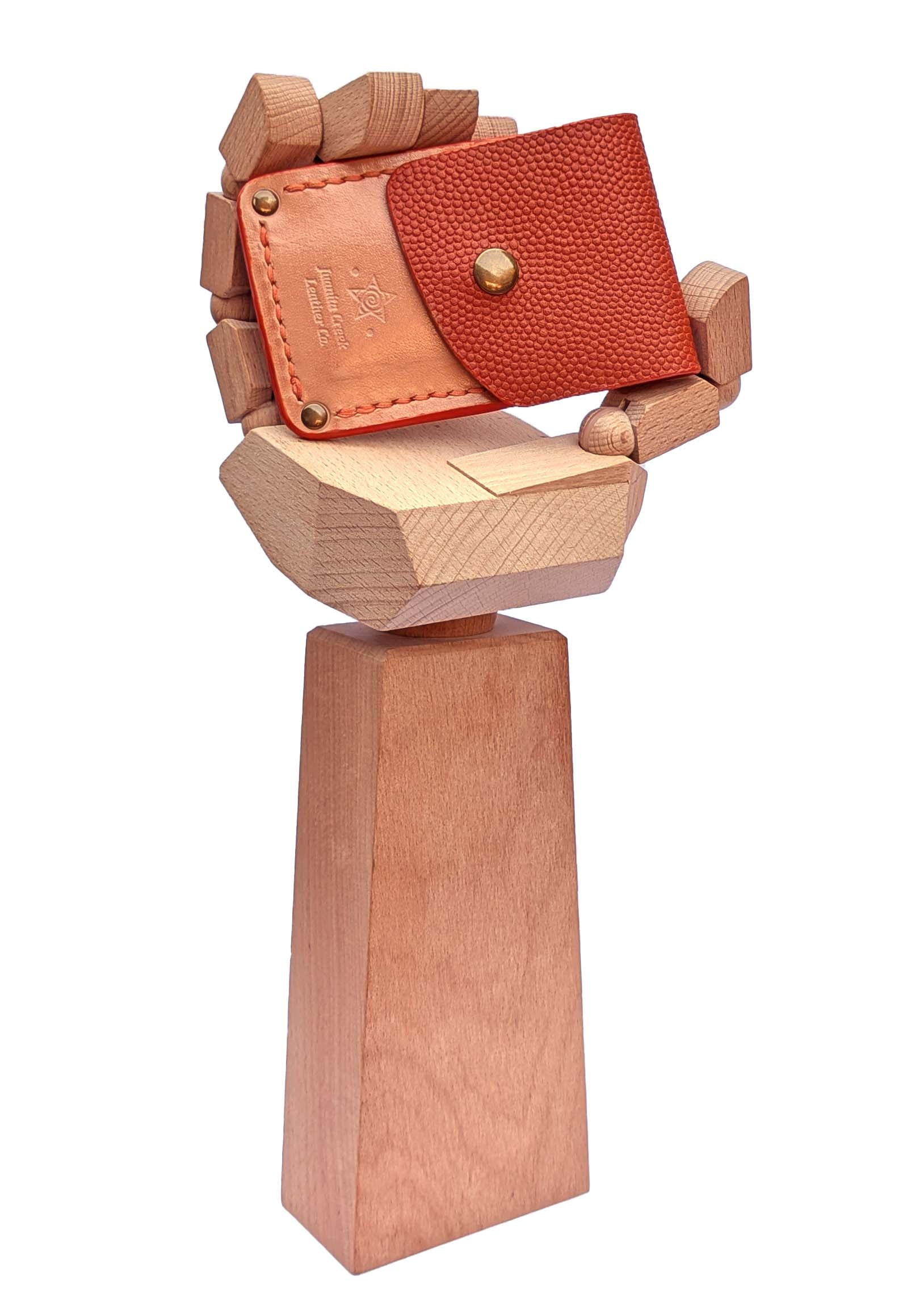 A small leather wallet or card holder in reddish-brown pebbled leather with tan stitched flap and brass-tone snap, displayed on a sculptural wooden stand against a plain white background.