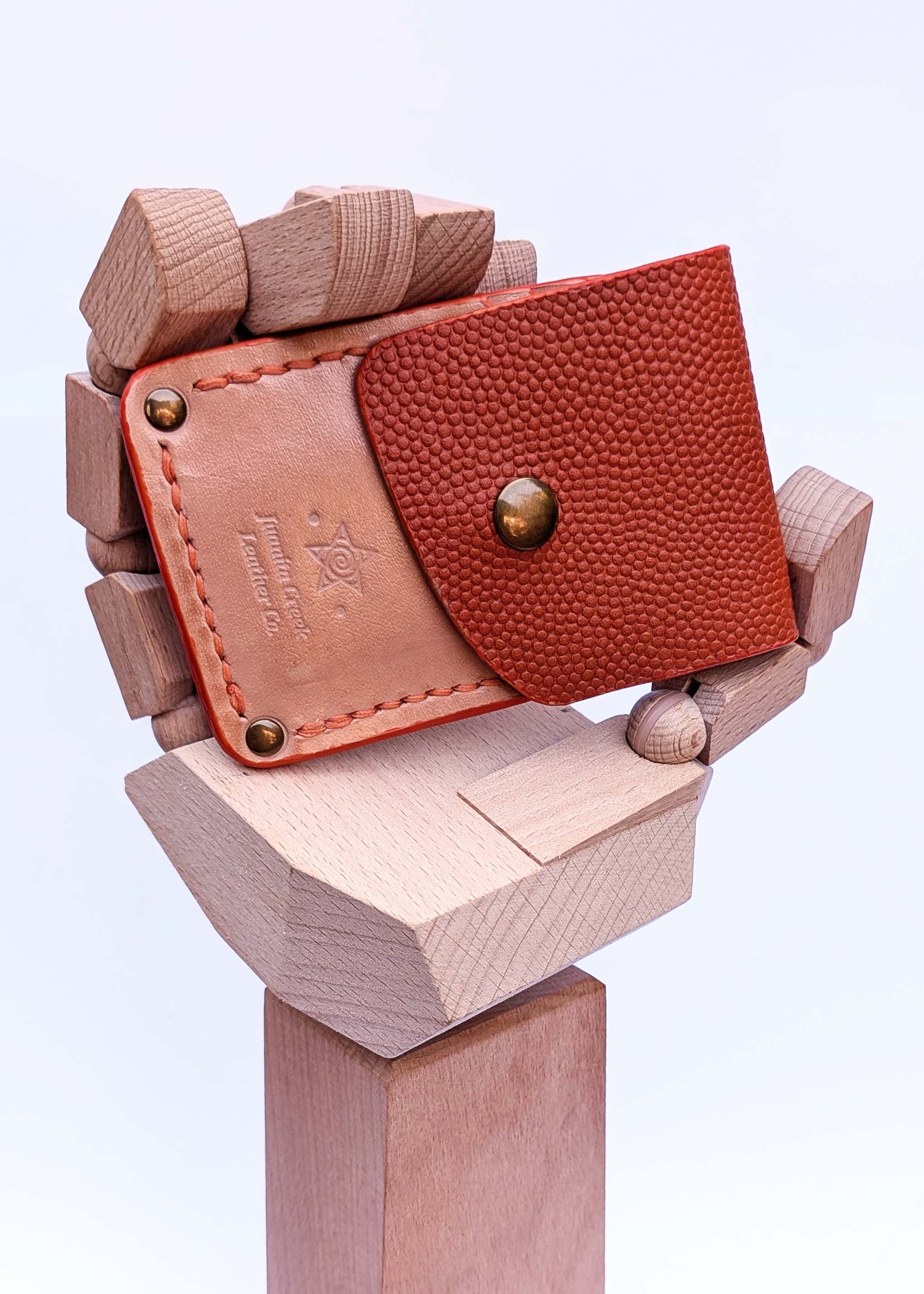 A small leather wallet or card holder with a textured reddish-brown pebbled flap and a tan leather back panel, displayed on a sculptural wooden stand against a clean white background.