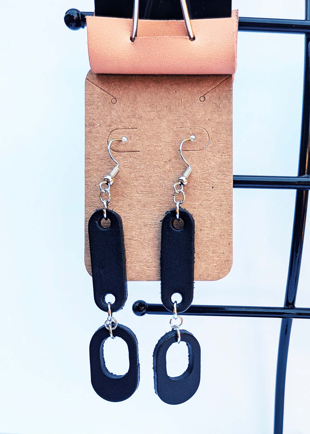 A pair of black leather dangle earrings with silver-tone hooks, displayed on a brown cardboard card held by a black metal stand against a white background.