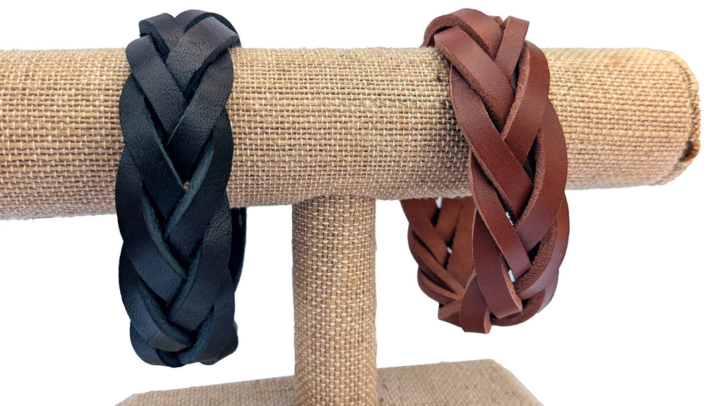 Two braided leather bracelets displayed on a burlap-covered jewelry stand, one in black and one in reddish brown, against a white background.