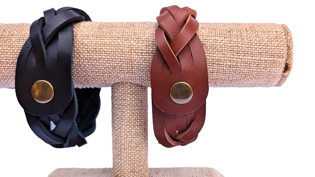 Two leather wrap bracelets displayed on a burlap-covered jewelry stand against a white background. One bracelet is black with a brass snap, and the other is reddish-brown with a matching brass snap.