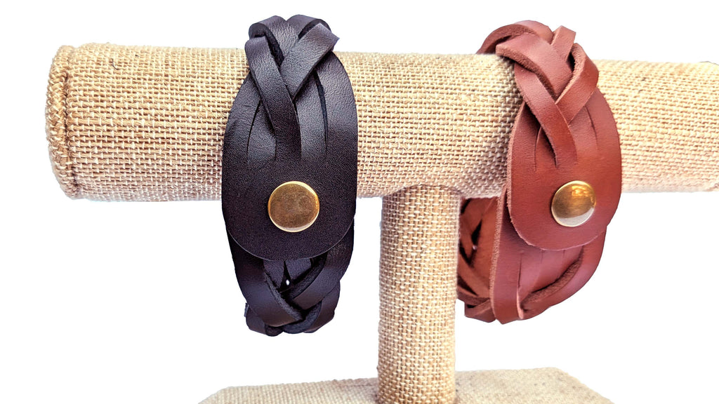 Two braided leather cuff bracelets displayed on a burlap-covered jewelry stand. One bracelet is black and the other is reddish-brown, both featuring a round brass snap button.